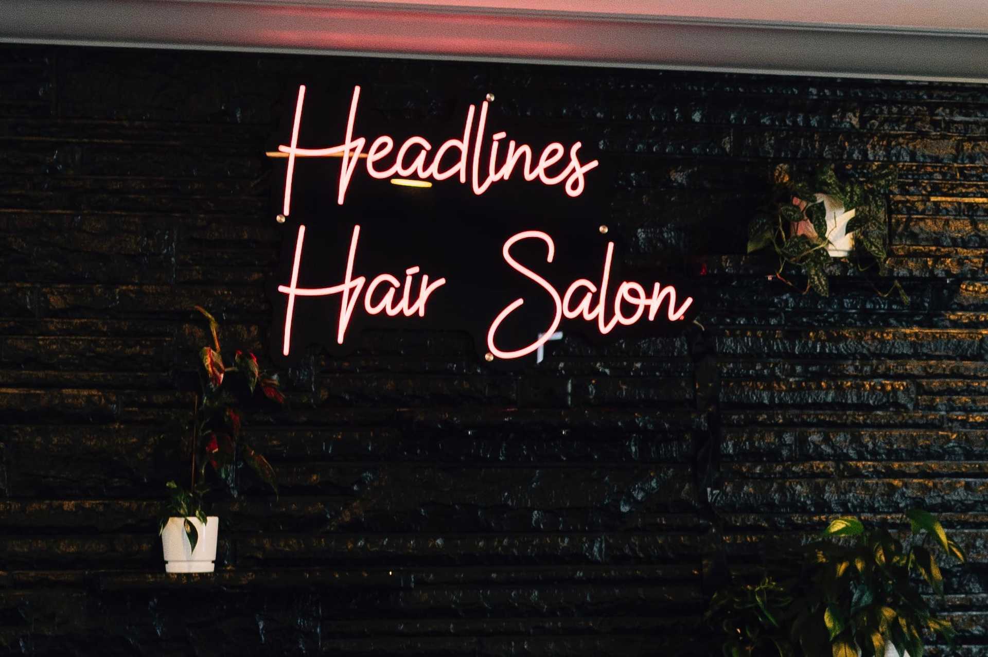 Headlines Hair Salon neon sign on a dark textured wall with plants.